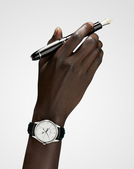 Main de homme tenant un stylo plume Montblanc et portant une montre Jaeger-LeCoultre sur fond blanc.